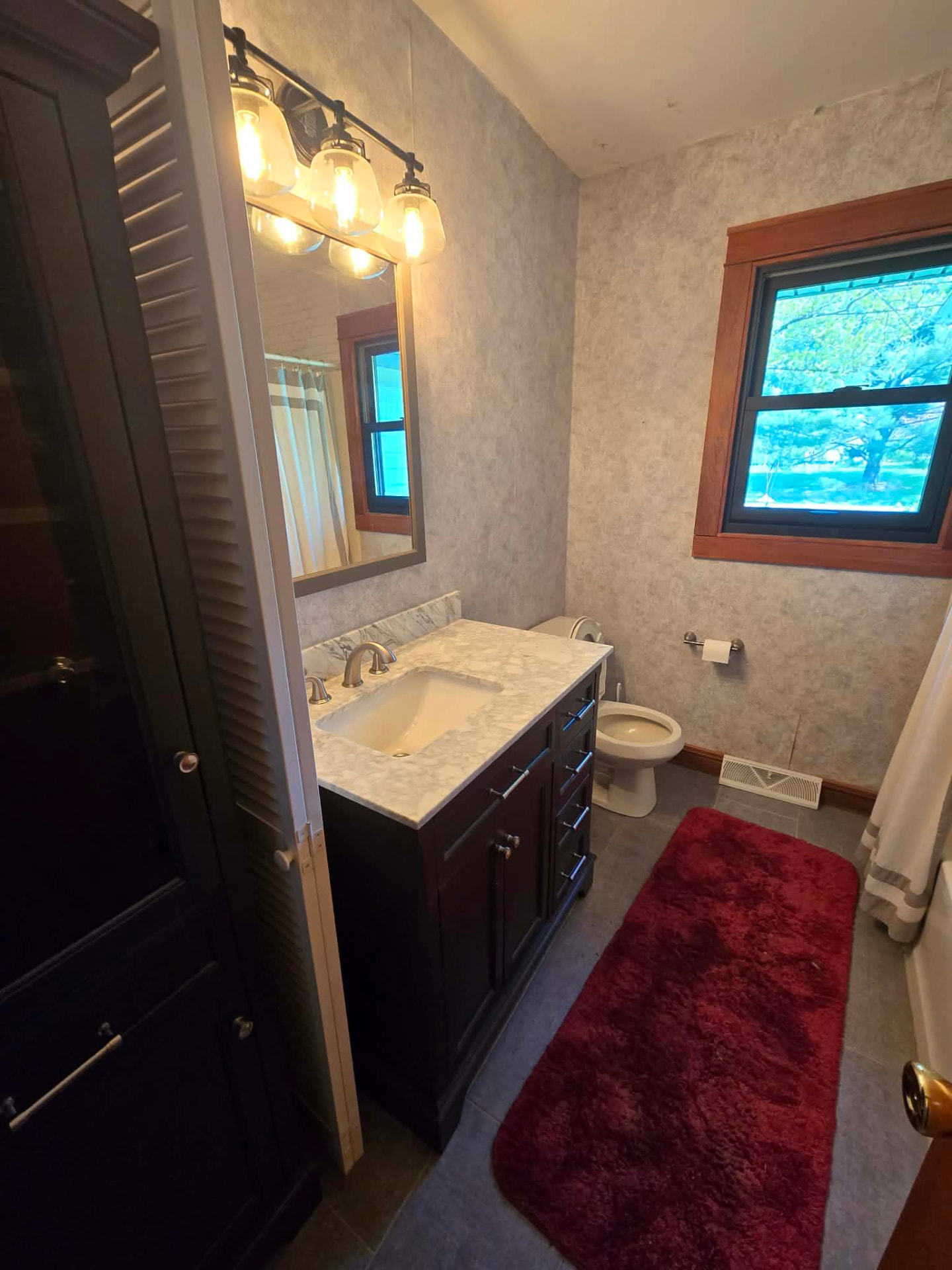 Before: dated hall bath with mottled wallpaper, dark traditional vanity, wood-trimmed window, and bifold linen closet doors