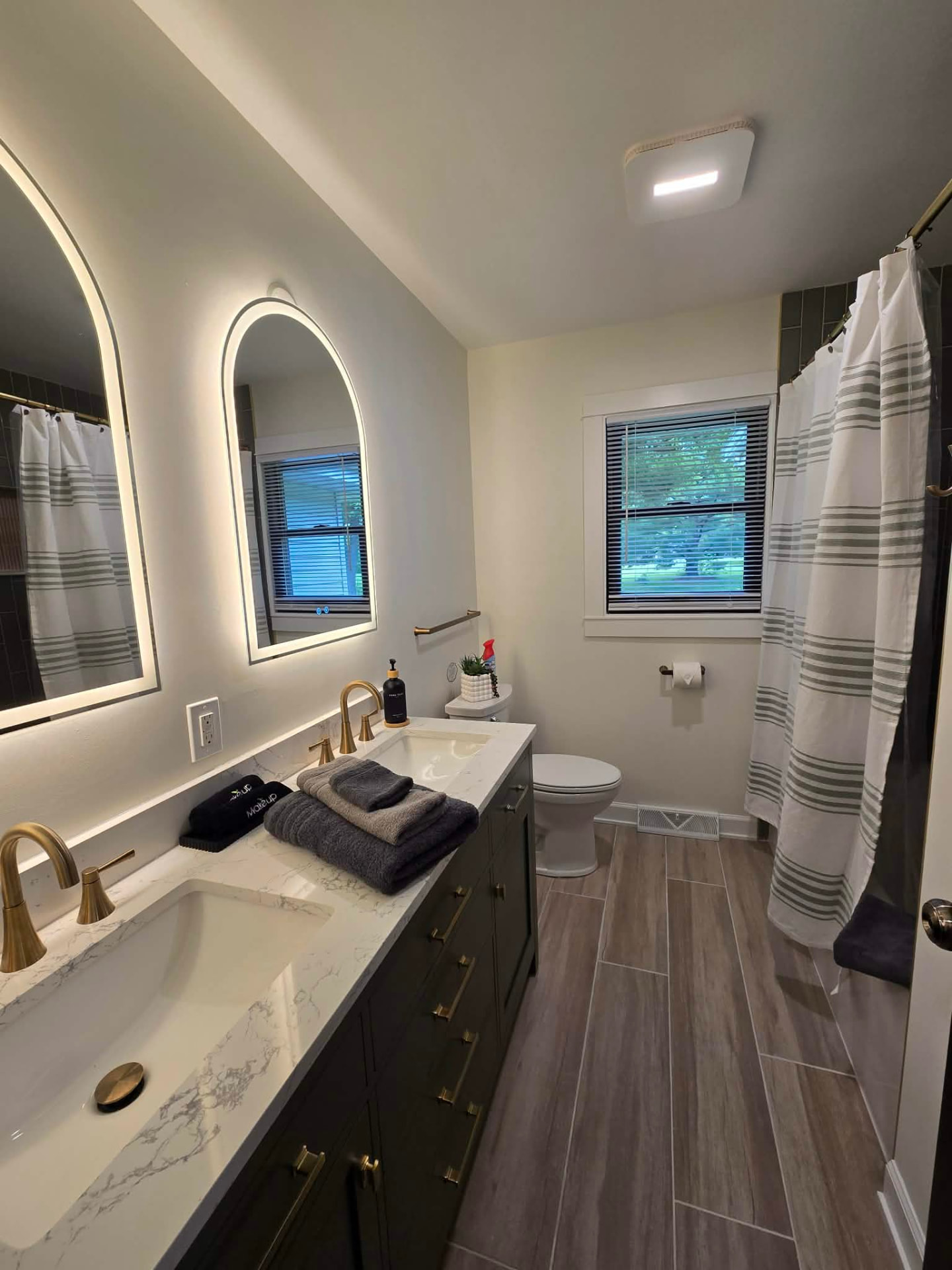 After: hall bath with a dark double vanity, quartz marble-look top, brass fixtures, arched LED mirrors, and wood-look plank tile floor