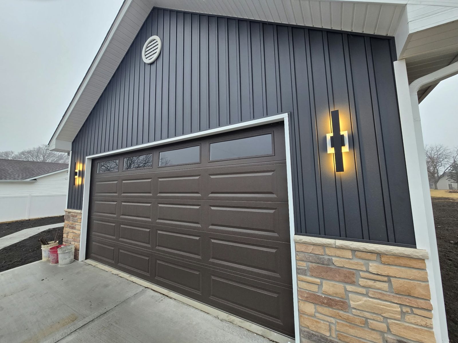 Attached garage with board-and-batten siding, stone base and modern wall lights