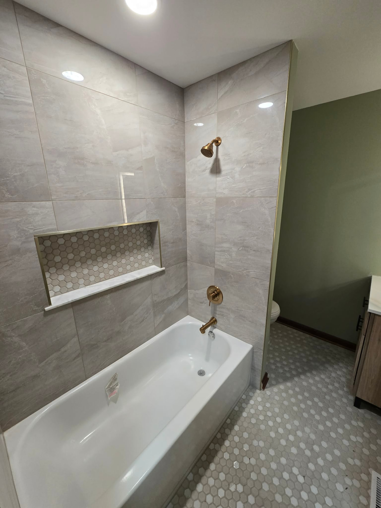After: guest bath with large-format marble-look tile tub surround, brass fixtures, hex mosaic niche and floor, and sage green walls